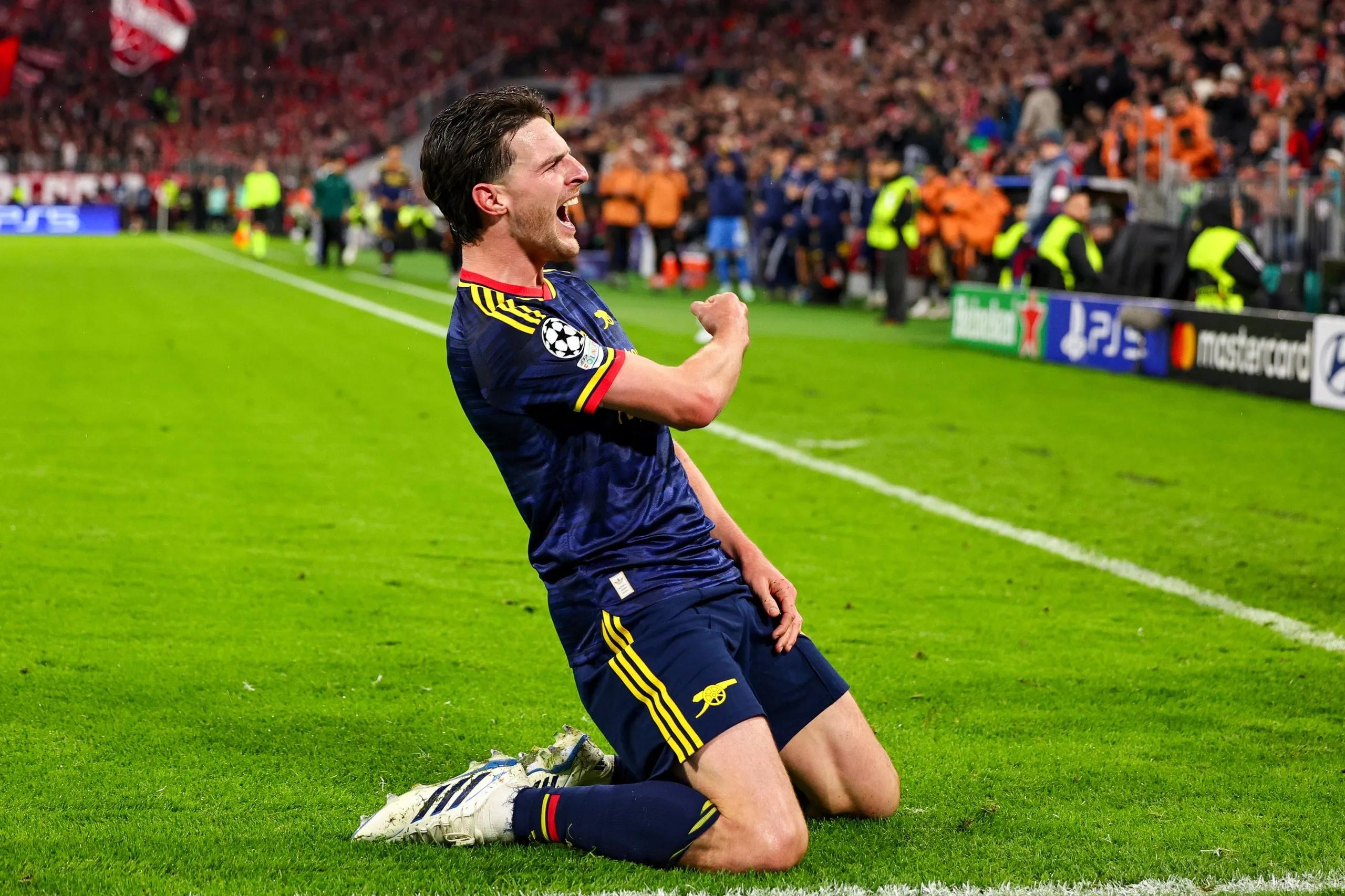 Declan Rice kneels on the pitch clenching his fist, celebrating a goal for Arsenal